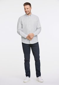 Camisa de botones con finas rayas verticales en gris claro y blanco, de manga larga, y con un bolsillo en el pecho, combinada con pantalones oscuros y zapatillas blancas.