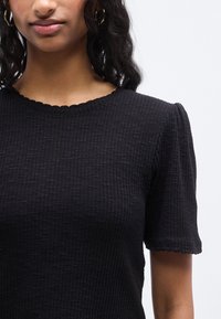 Μαύρο τοπ με υφή και κοντά μανίκια με κυματιστό τελείωμα. Κατασκευασμένο από ριμπ ύφασμα με λεπτά υφασμένα σ χέδια. Στρογγυλή λαιμόκοψη.