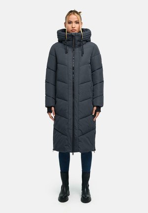 Femme portant un long manteau d’hiver matelassé gris foncé avec une grande capuche, un jean bleu et des bottes noires, debout devant un fond clair uni.