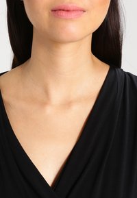 Sort indpakningsbluse med V-hals, glat tekstur og blødt stof. Nærbillede af halsen og den øvre brystområde.
