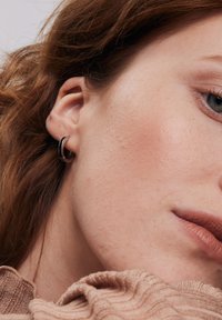 Czarna, okrągła kolczyk w kształcie obręczy, z metalicznym wykończeniem, przymocowany do ucha modelki. Miękka tekstura skóry i lekki makijaż, widoczny neutralny sweter.