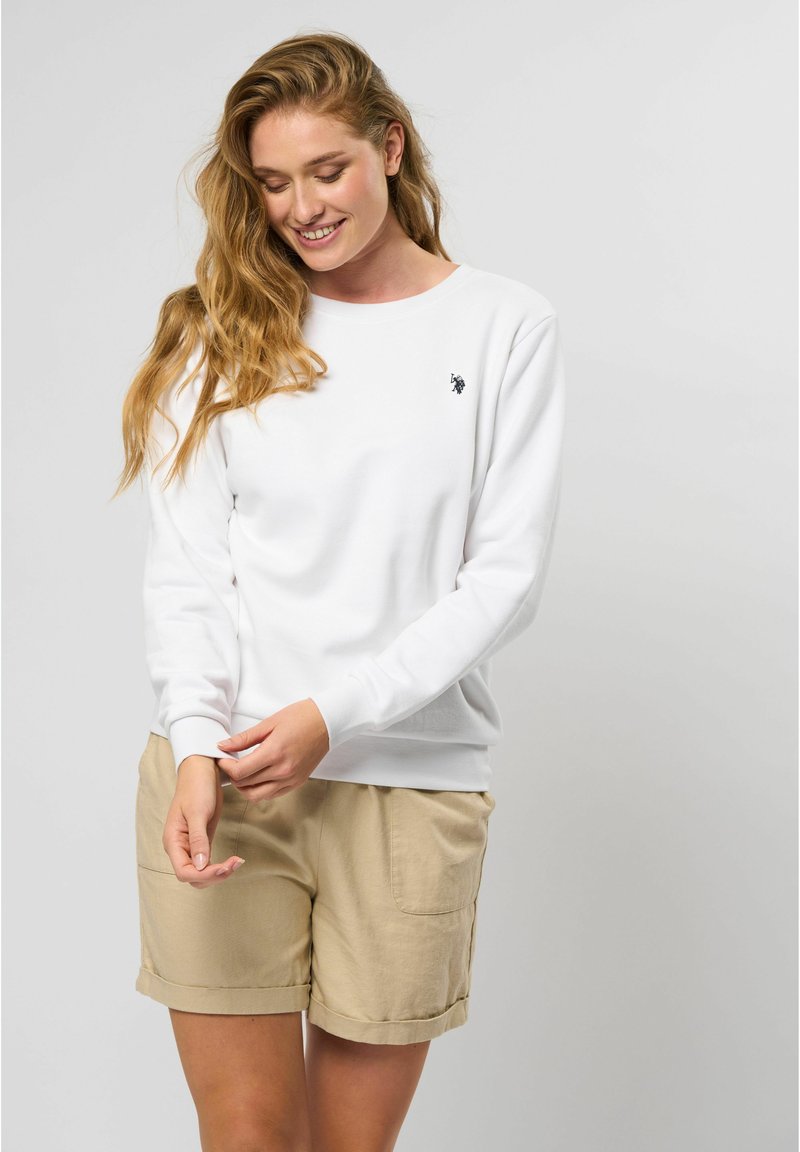 Balta jaka ar garām piedurknēm, apaļu izgriezumu un mazu melnu logotipu uz krūtīm, apvienota ar krēmkrāsas šortiem ar sānu kabatām.