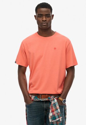 Krótki rękaw, t-shirt z okrągłym dekoltem w kolorze koralowym, wykonany z bawełny, z małym logo na piersi, zestawiony z niebieskimi dżinsami i kraciastą koszulą zawiązaną w talii.