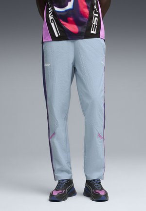 Pantalon de sport bleu clair texturé avec des bandes latérales noires et roses, associé à des chaussures de course noires et roses, porté par un mannequin debout.