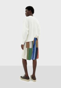 Weiße Langarmbluse kombiniert mit mehrfarbigen Patchwork-Shorts, die grüne, blaue, braune und rote Streifen aufweisen, sowie dunklen Schuhen mit cremfarbenen Sohlen.