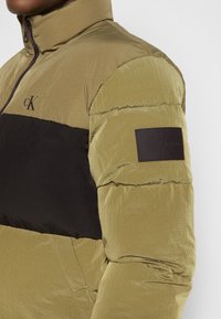 Pufová bunda s khaki vrchní částí a černou spodní částí. Obsahuje vysoký límec, zipové zapínání a strukturovanou tkaninu s koženou značkovou nášivkou.