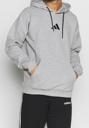Mężczyzna w jasnoszarej bluzie Adidas z kapturem z czarnymi sznureczkami oraz czarnych spodniach dresowych Adidas z białymi paskami, ręce w kieszeniach bluzy.