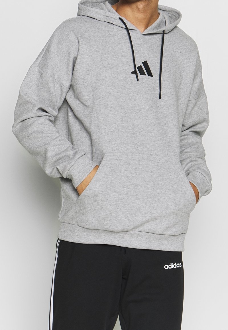 Homme portant un sweat à capuche Adidas gris clair avec des cordons noirs et un pantalon de survêtement Adidas noir avec des rayures blanches, les mains dans les poches du sweat.