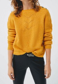 Femme aux cheveux bouclés portant un pull en tricot jaune moutarde avec des boutons sur les épaules et un pantalon noir avec une ceinture.