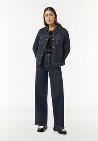Dunkelblaue Denimjacke mit zwei Brusttaschen und Kontrastnähten, kombiniert mit hoch taillierten, weit geschnittenen dunklen Jeans und schwarzen Schuhen.