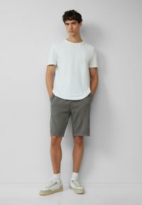 Witte crewneck t-shirt, grijze katoen shorts en lichte sneakers met groene accenten. Model staat tegen een effen achtergrond.