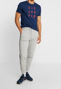 Marineblå T-shirt med rødt "STOCKHOLM"-tryk, kombineret med lysegrå joggingbukser med sidestriber og elastik i taljen. Sorte sneakers.