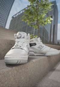 White high-top sneakers with black accents, featuring a smooth leather texture, prominent logo, and padded collar. Positioned on stone steps.