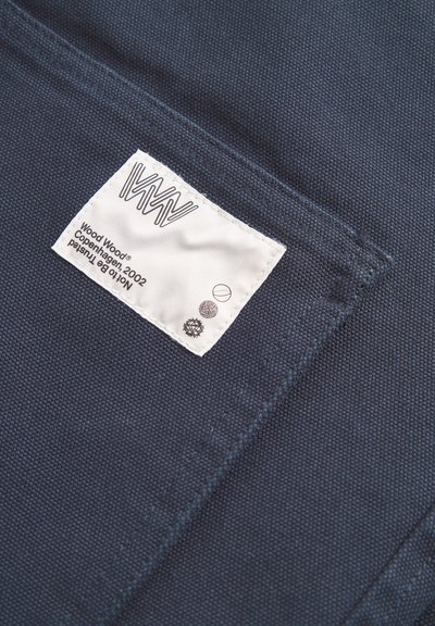 Material de culoare albastru închis cu o țesătură texturată, prezentând un eticheta de brand albă cosută deasupra cu simboluri negre și colorate.