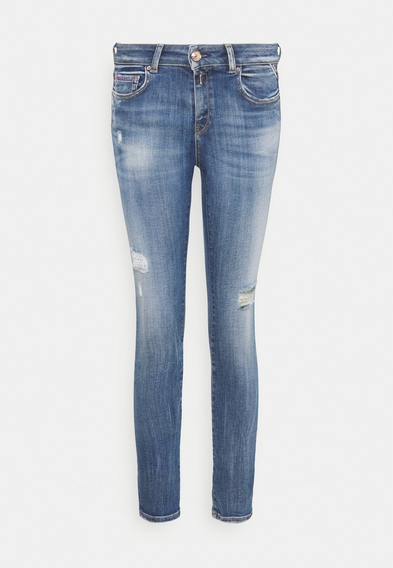 Jean slim bleu avec légère décoloration et petites zones usées sur les cuisses, bouton avant et passants de ceinture visibles.