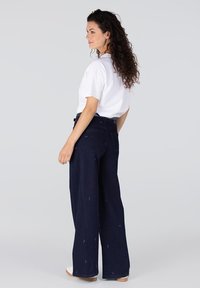 Donkerblauwe wijde jeans met subtiele borduursels, gecombineerd met een witte shirt met korte mouwen. Model wordt van achteren getoond, staande tegen een effen achtergrond.