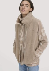 Jonge vrouw in een beige bloemenfleecejack, gelaagd met een beige ritsvest, staand met haar handen in de zakken tegen een effen achtergrond.