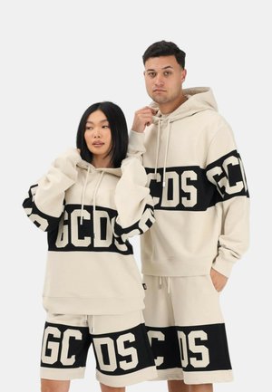 Man en vrouw dragen bijpassende beige hoodies en shorts met grote zwarte GCDS-logo's over de borst en benen, poserend tegen een witte achtergrond.