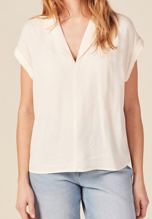 Woman wearing a short-sleeve, white V-neck blouse paired with light blue jeans against neutral background.