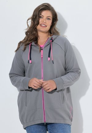 Donna che indossa una felpa grigia con zip e dettagli rosa e jeans blu, in piedi contro un muro bianco, sorridente con le mani giunte.
