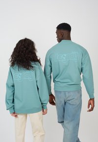 Turkise gensere med rund hals og mansjetterte ermer, med lys blå "Legacy" tekst på baksiden. Uformell, komfortabel design.