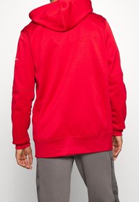 Sweat à capuche rouge avec une finition texturée, doté d'une capuche, de manches longues et de poignets côtelés. Le design comprend des coutures subtiles et un accent logo.