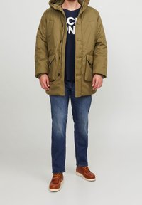 Oliivinvihreä eristetty takki, jossa on huppu, etunapit ja kaksi suurta rintataskua, yhdistettynä sinisiin farkkuihin ja ruskeisiin kenkiin.