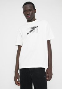 Biały bawełniany t-shirt z czarnym nadrukiem graficznym przedstawiającym but. Okrągły dekolt, krótkie rękawy, luźny krój, w zestawieniu z czarnymi jeansami.