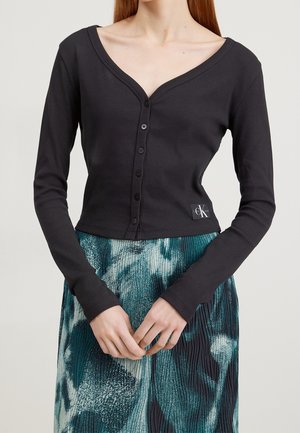 Woman wearing a black buttoned long-sleeve V-neck top and a pleated skirt with blue, teal, and white abstract patterns.