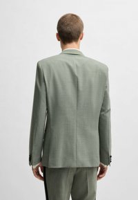 Blazer verde chiaro realizzato in tessuto texturizzato. Presenta un rever a punta, una fessura posteriore e accenti di bottoni neri sulle maniche. Cuciture laterali visibili.