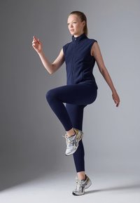 Temno modra brezrokavnik in usklajene legice iz raztegljive tkanine, kombinirane s srebrnimi športnimi čevlji, z motivom v dinamičnem položaju.