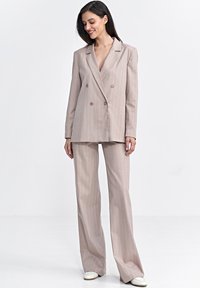 Jasno beżowa, dwurzędowa marynarka z pionowymi paskami, zestawiona z pasującymi spodniami o wysokim stanie i szerokich nogawkach. Posiada klapy i dwa guziki.