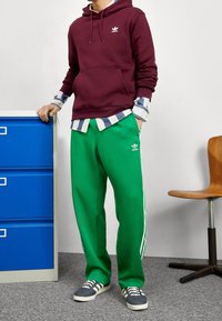 Sudadera burdeos con bolsillo tipo canguro, pantalones de chándal verdes con rayas blancas y zapatos deportivos azul y blanco. Modelo de pie contra un armario azul.