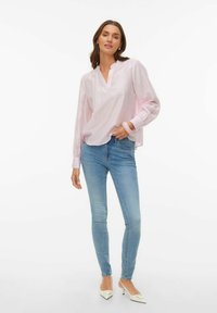 Šviesiai rožinė palaidinė su V formos iškirpte ir raukšlėtomis ilgomis rankovėmis, derinama su vidutinio mėlynumo lieknomis džinsinėmis kelnėmis ir smailiais baltais aukštakulniais.