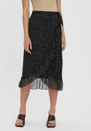 Femme portant une jupe portefeuille midi noire à pois blancs avec un ourlet à volants, associée à des sandales noires à talons ouverts.