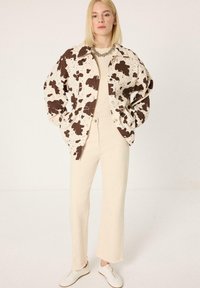 Γυναίκα φοράει oversized κρεμ μπουφάν με καφέ μοτίβο αγελάδας, κρεμ παντελόνι, λευκά αθλητικά παπούτσια και χοντρό ασημένιο κολιέ, στέκεται με τα χέρια στις τσέπες.