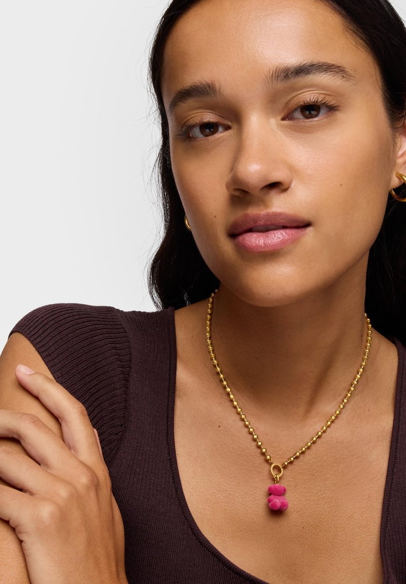 Collana di perle d'oro con ciondolo rosa peloso. Modella che indossa una maglia scura a coste. La pelle liscia e il trucco naturale valorizzano i gioielli.