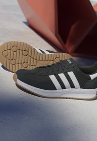Zapatilla negra Adidas con rayas blancas y suela de goma, posicionada sobre hormigón con una escultura geométrica de color óxido en el fondo.
