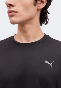 Νεαρό άτομο που φοράει μαύρο T-shirt Puma με εμφανές γκρίζο λογότυπο Puma στο αριστερό στήθος, σε απλό ανοιχτό φόντο.