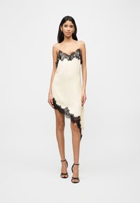 Γυναίκα που φοράει ασύμμετρο φόρεμα μίνι από σατέν σε εκρού απόχρωση με μαύρη δαντέλα και μαύρα ψηλοτάκουνα σανδάλια, στέκεται μπροστά σε απλό φόντο.