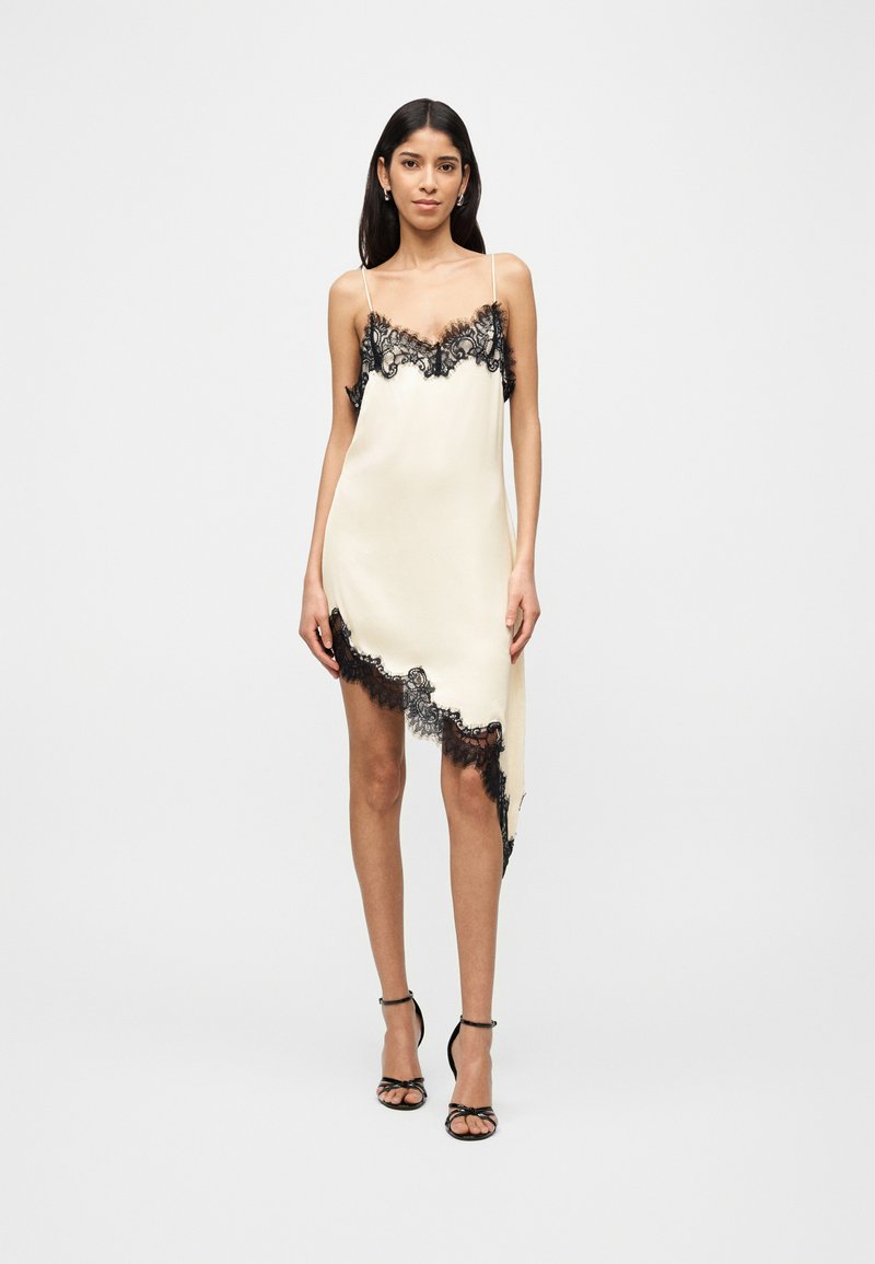 Γυναίκα που φοράει ασύμμετρο φόρεμα μίνι από σατέν σε εκρού απόχρωση με μαύρη δαντέλα και μαύρα ψηλοτάκουνα σανδάλια, στέκεται μπροστά σε απλό φόντο.