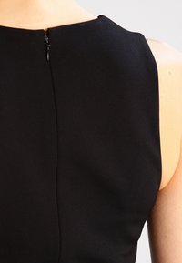 Vestido negro sin mangas que presenta una tela suave y mate con un discreto cierre en la parte posterior y un ligero detalle de costura en la espalda.