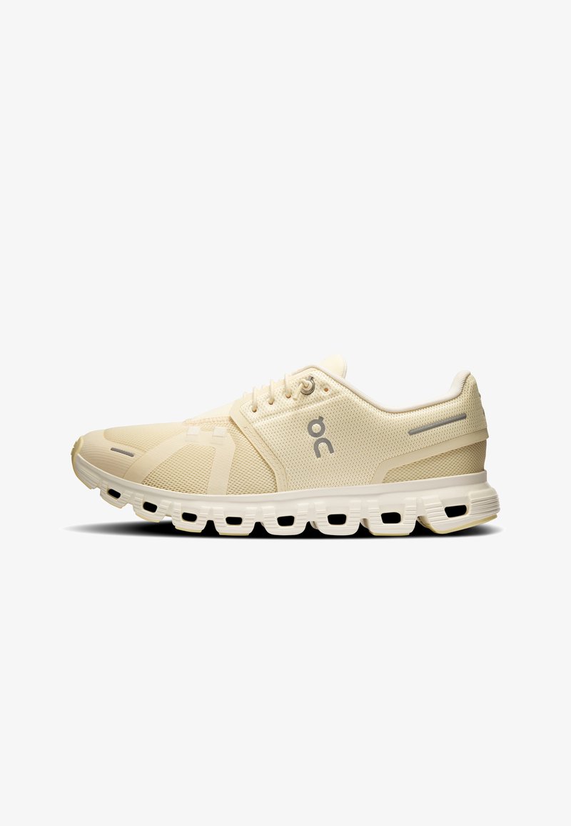 Beiger Sportschuh mit Mesh-Obermaterial, Schnürverschluss, gepolsterter segmentierter Sohle und reflektierenden Details an Ferse und Zehenbereich.