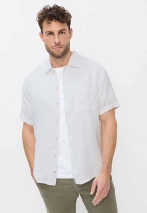 Mann mit lockigem Haar, der ein weißes kurzärmeliges Hemd über einem weißen T-Shirt und olivgrünen Hosen trägt, steht vor einem schlichten Hintergrund.