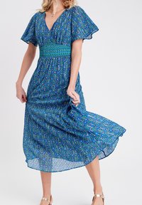 Robe maxi bleue à motifs avec de courtes manches volantées, une taille smockée et un ourlet fluide. Présente un design géométrique en bleu foncé et bleu clair.