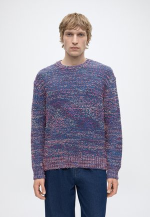 Homme blond portant un pull en tricot texturé bleu et rose à manches longues et un jean bleu foncé, debout devant un fond clair uni.