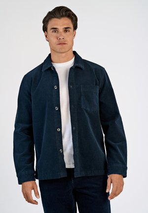 Man draagt donkerblauw corduroy overhemd over een wit T-shirt en staat tegen een effen lichte achtergrond.