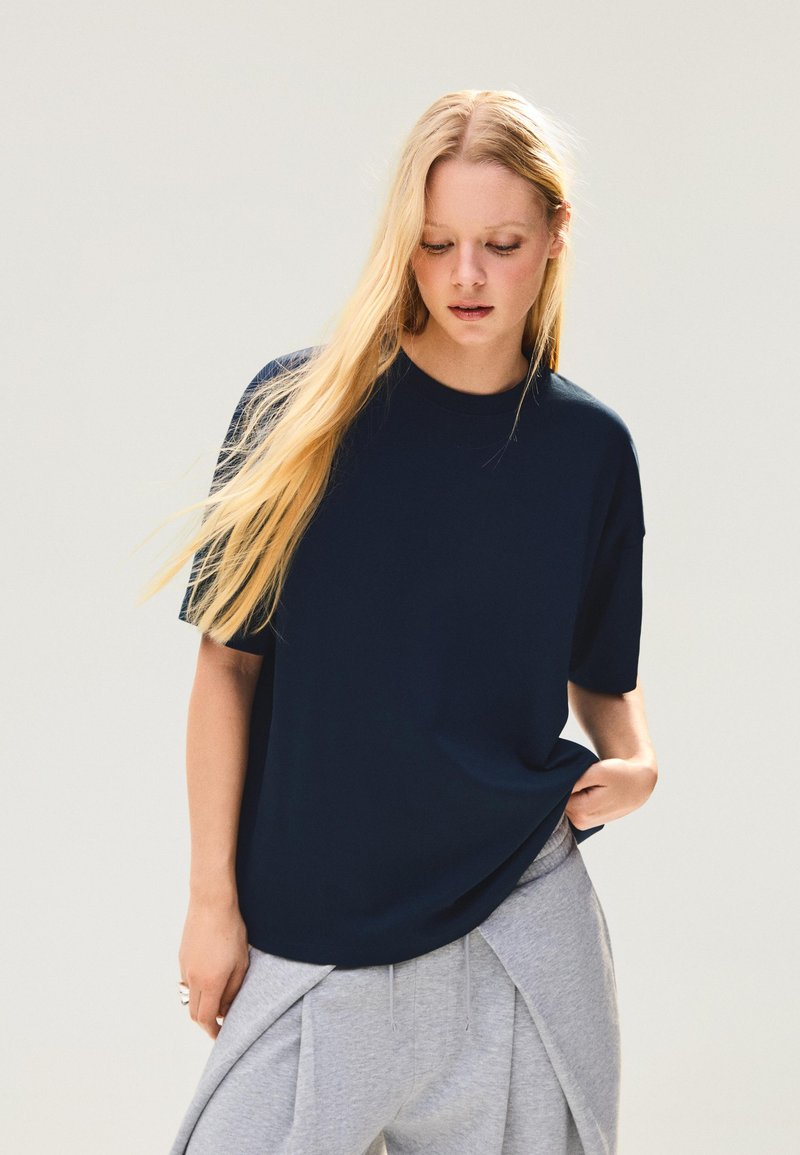 Jeune femme aux longs cheveux blonds portant un t-shirt ample bleu marine et un pantalon gris clair, debout devant un fond clair uni.
