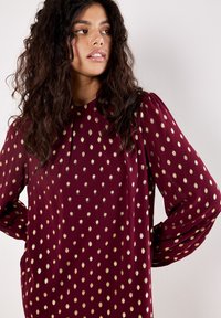 Donna con capelli ricci che indossa un vestito a maniche lunghe color bordeaux con pois dorati, che guarda di lato su uno sfondo bianco semplice.