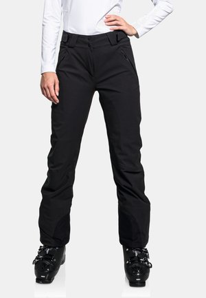 Vrouw draagt zwarte skibroek en zwarte skistappers, staat met één hand op haar heup en de andere ontspannen langs haar zij.